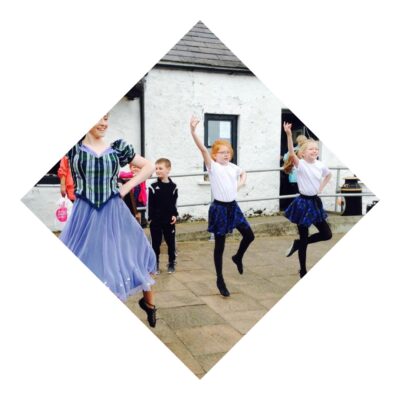Three young dancers perform outdoors in traditional Scottish attire, with two girls in blue tartan skirts and black tights mid-step and a woman in a flowing purple skirt leading.
