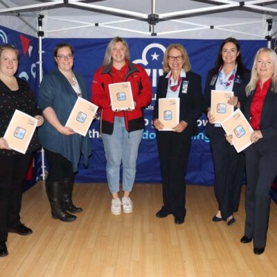 Six women stand inside a tent decorated with Girlguiding logos, each holding a certificate folder and smiling at the camera.