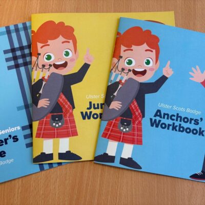 Three colourful Ulster Scots Badge workbooks with blue and yellow covers. The books feature cartoon characters in traditional Scottish attire.